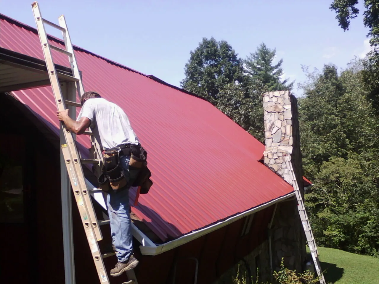 Roofer on a ladder beside a red metal roof and stone chimney — Soffit & Fascia Repair by Roofing Brothers in Rumson